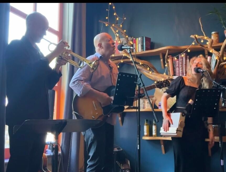 A trumpet player, guitarist and singer performing together beside bookshelves